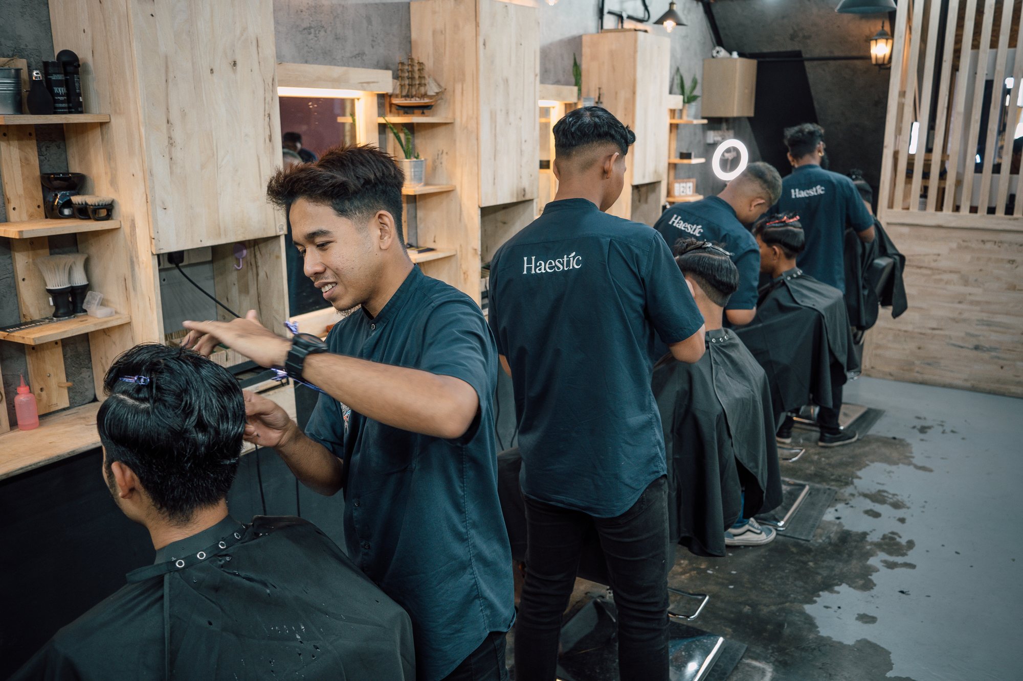 Haestic barbers in navy shirts at work along a row of chairs in a warm, wood-panelled barbershop