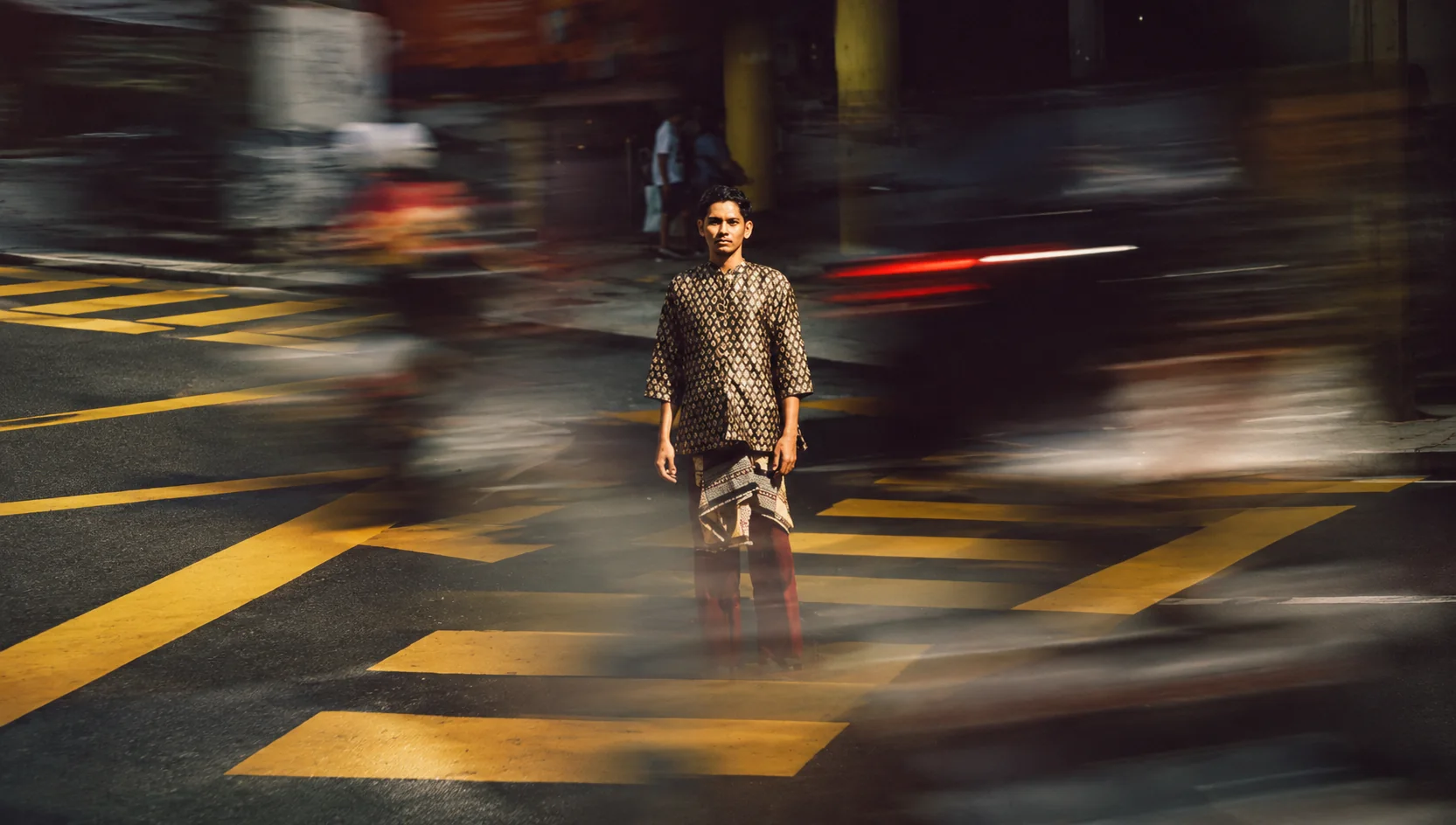 A BudiMan in a songket-print shirt standing on a zebra crossing as traffic blurs past
