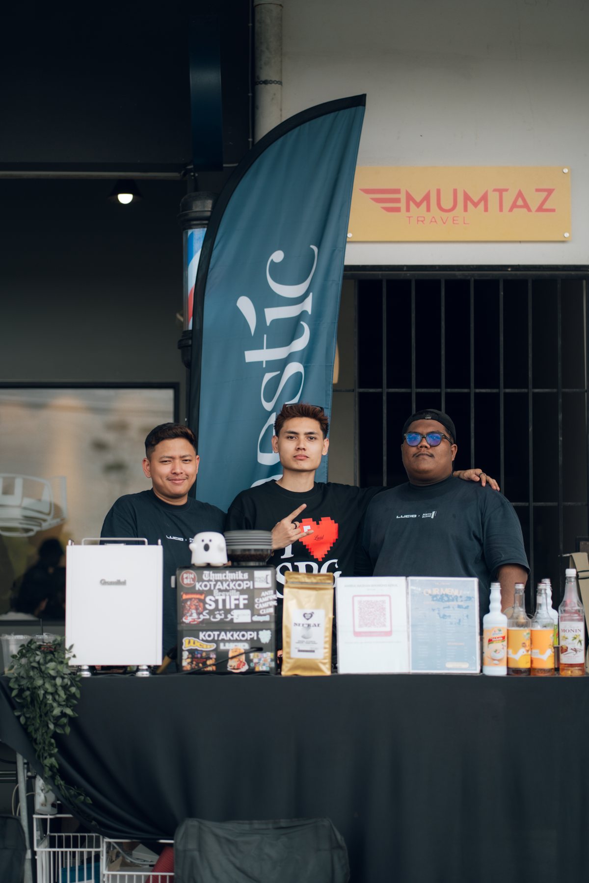 Three members of the Lucid team smiling behind a coffee stall, with a tall Haestic flag banner behind them