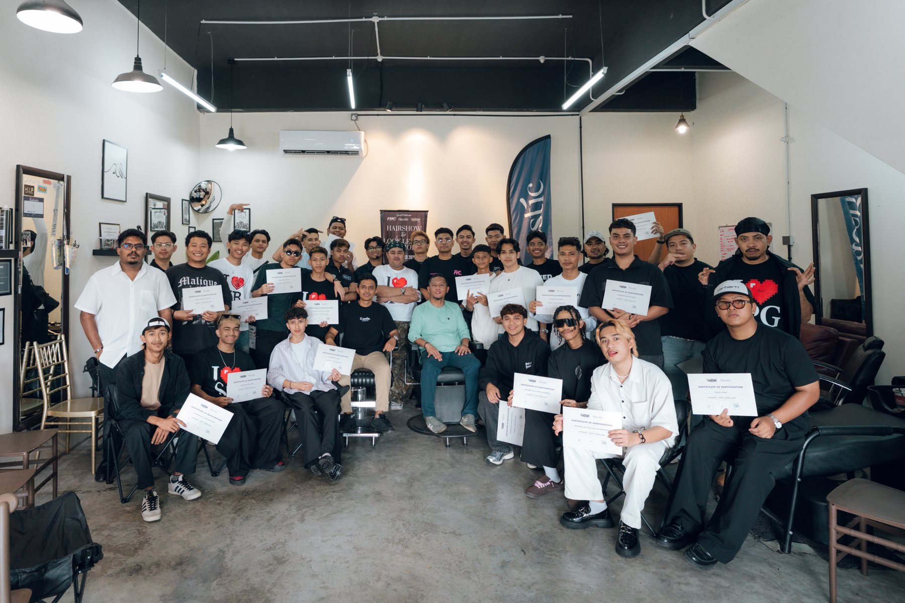 A large group of barbers holding certificates of participation inside a barbershop, with a Haestic flag banner behind them